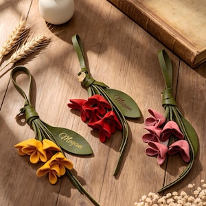 Könnte beinhalten: Drei Blumen-Schlüsselanhänger aus Leder mit gelben, roten und rosafarbenen Blütenblättern an grünen Stielen und Blättern. Jeder Anhänger hat eine Schlaufe und ein blattförmiges Etikett, das mit dem Namen Megan in Schreibschrift graviert ist.