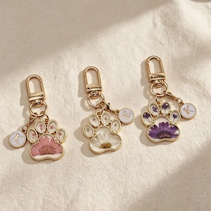 Peut inclure: Trois porte-clés en métal doré en forme de patte d'animal, contenant des fleurs séchées roses, blanches et violettes. Chaque porte-clé est doté d'un fermoir mousqueton et d'une petite breloque circulaire avec les lettres K, E ou K en blanc.