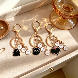 May include: Three gold-toned metal keychains featuring lobster clasps and split rings. Each keychain includes a unique enamel charm: a black ghost, a black horned creature, and a black ghost with a red mouth, accompanied by small white letter charms.