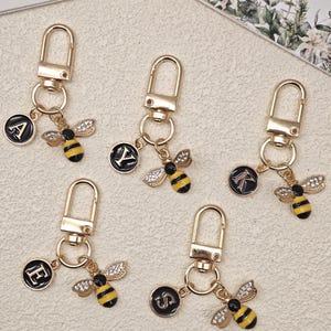 May include: Five gold-toned metal keychains, each featuring a lobster clasp, a circular black charm with a white letter, and a bee charm. The bee charms have yellow and black striped bodies with clear crystal-encrusted wings. The letters are A, Y, K, E, and S.