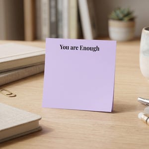 May include: A square light purple sticky note standing upright on a wooden surface. The note features the text "You are Enough" printed in a simple black serif font at the top centre, leaving the rest of the paper blank for writing notes.