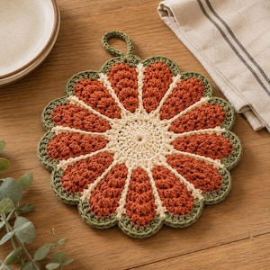 May include: A round, crocheted pot holder featuring a central cream-coloured circle with radiating segments in burnt orange, bordered by a scalloped edge in sage green. The design includes a small crocheted loop at the top for hanging purposes.