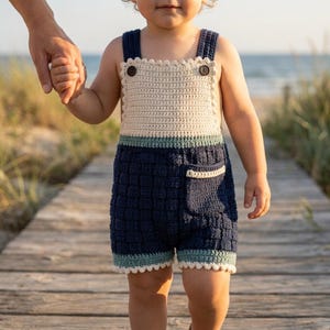 May include: A crochet romper featuring a cream-colored bib with two dark brown buttons, a navy blue textured lower section with a small pocket, and light green trim at the waist and leg openings. The edges are finished with a scalloped crochet border.