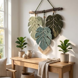 May include: A wall hanging featuring three crocheted monstera leaves in varying shades of green, suspended from a dark wooden branch with a hanging cord. The leaves are detailed with central stitching and the bottom leaf has a cream-coloured fringe.