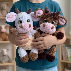 Op de afbeelding: Twee handgemaakte gehaakte knuffelkoeien. De ene koe is wit met bruine vlekken, bruine hoorns en een roze snuit. De andere koe is bruin met een roze snuit, bruine hoorns en donkerbruine hoeven. Beide hebben grote, ronde, glanzende ogen met wimpers.