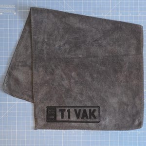May include: A dark grey microfibre towel featuring a rectangular black embroidered patch in the bottom corner. The patch displays the text T1 VAK alongside a small UK flag icon. The fabric has a soft, textured surface with finished edges.