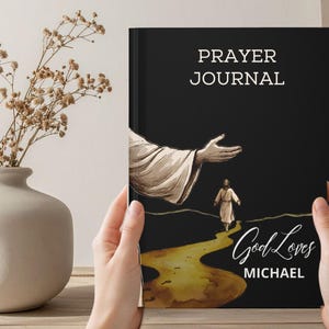 Op de afbeelding: Een zwart gebedsdagboek met witte tekst met de tekst PRAYER JOURNAL en GOD LOVES MICHAEL. De omslag bevat een illustratie van een grote hand die reikt naar een klein figuur dat op een gouden pad loopt tegen een donkere achtergrond.