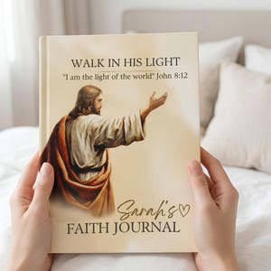 Op de afbeelding: Een dagboek met harde kaft in crèmekleur met een illustratie van een figuur in gewaden met een uitgestoken hand. De tekst luidt WALK IN HIS LIGHT, I am the light of the world John 8:12, Sarah's FAITH JOURNAL met een hartpictogram.
