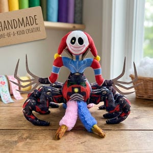 May include: A plush creature with a white face, red jester-style hat, and a wide stitched smile. It has a blue and red torso, long pointed fingers, and multiple dark patterned legs. Two fuzzy pink and blue appendages extend from the center.