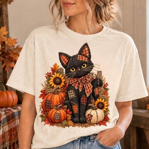 May include: A cream-colored short-sleeve t-shirt featuring a graphic of a black cat with yellow eyes, wearing a plaid bow tie. The cat is surrounded by orange pumpkins, sunflowers, and autumn leaves, with patches of plaid fabric on its body.