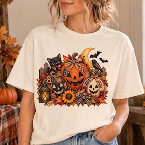 May include: A cream-coloured short-sleeve t-shirt featuring a graphic print of three jack-o'-lanterns, a black cat, a crow, a crescent moon, bats, autumn leaves, a sunflower, and a lantern, all arranged in a festive autumn-themed composition.