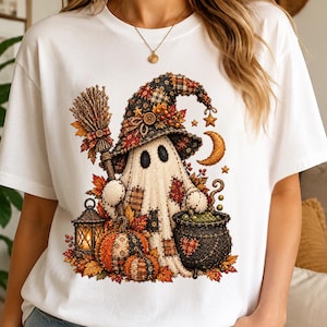 May include: A white t-shirt featuring a graphic of a ghost wearing a patchwork witch hat, holding a broomstick. The design includes two pumpkins, a lit lantern, a cauldron, autumn leaves, a crescent moon, and small stars in orange and brown.
