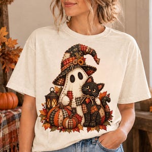 May include: A cream-colored short-sleeve t-shirt featuring a graphic print of a ghost wearing a plaid patchwork witch hat, standing next to a black cat with plaid accents, surrounded by pumpkins, autumn leaves, and a small lit lantern.