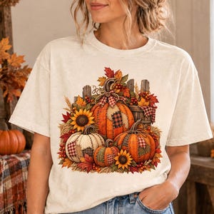Peut inclure: Un t-shirt à manches courtes de couleur crème avec une impression graphique centrale représentant trois citrouilles orange, blanches et vertes, agrémentées de tournesols, de feuilles d'automne, de baies et d'une clôture en bois.
