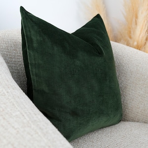 May include: A square throw pillow featuring a deep forest green velvet fabric cover. The pillow has a soft, plush texture with a subtle sheen and a clean, knife-edge finish along the seams, presenting a solid, uniform color throughout the surface.