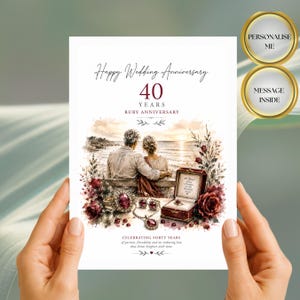 以下が含まれることがあります： 40周年ルビー婚式用の白いグリーティングカード。海辺に座るカップル、ルビーのジュエリー、赤いバラの水彩画が描かれています。テキストには「Happy Wedding Anniversary」「40 YEARS」「RUBY ANNIVERSARY」「CELEBRATING FORTY YEARS of passion, friendship and an enduring love that shines brighter with time」と記されています。