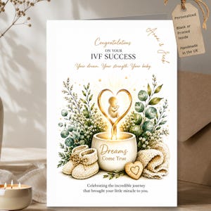 Op de afbeelding: Een witte wenskaart met de tekst Congratulations ON YOUR IVF SUCCESS, Your dream. Your strength. Your baby. en Dreams Come True. De illustratie toont een baby in een gouden hart boven een kom, met gebreide babyslofjes en groen blad.