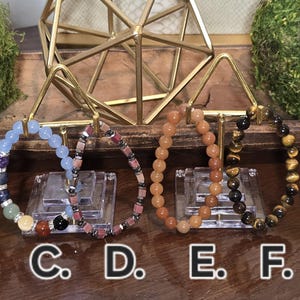 May include: Eight beaded bracelets labeled A through H, displayed on individual clear stands. The bracelets feature various natural stone beads in colors including brown, blue, purple, green, orange, black, and pink, arranged in circular patterns.