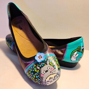 May include: A pair of brown leather flats featuring hand-painted illustrations of Totoro characters and blue flowers on the toe and heel. The shoes have a black trim, a light blue flower embellishment on the vamp, and a tan interior lining.