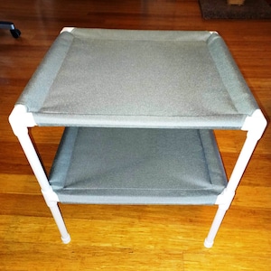 May include: Two-tiered cat bed made of white PVC pipe and gray fabric. The bed is elevated off the ground and has a simple, modern design.