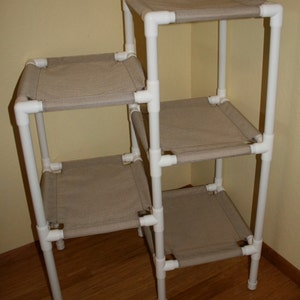 May include: A white PVC pipe cat tree with three platforms covered in beige fabric. The cat tree is in a corner of a room with wood flooring.