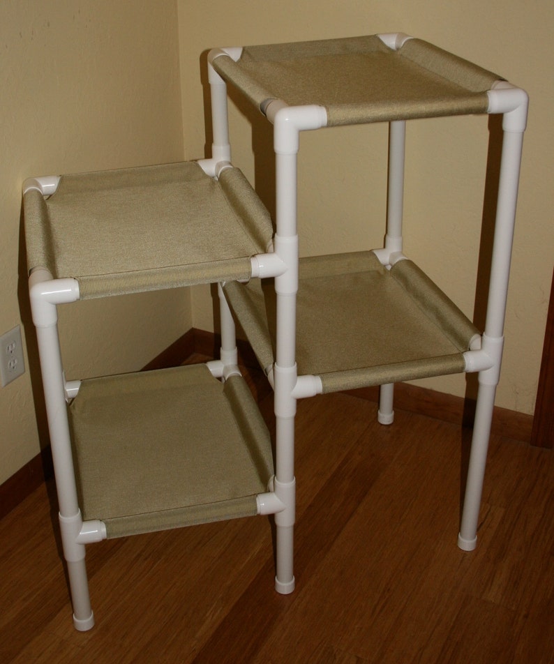 Pu&ograve; includere: Un albero per gatti a tre livelli realizzato in tubo di PVC bianco con ripiani rivestiti in tessuto beige. L'albero per gatti &egrave; assemblato in un angolo di una stanza con pavimento in legno.