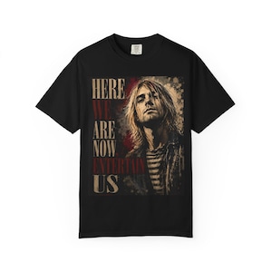 Peut inclure: Un t-shirt noir à manches courtes avec une impression graphique représentant une personne aux cheveux longs. Le texte "HERE WE ARE NOW, ENTERTAIN US" est imprimé dans une police vieillie sur la gauche, en rouge et beige.