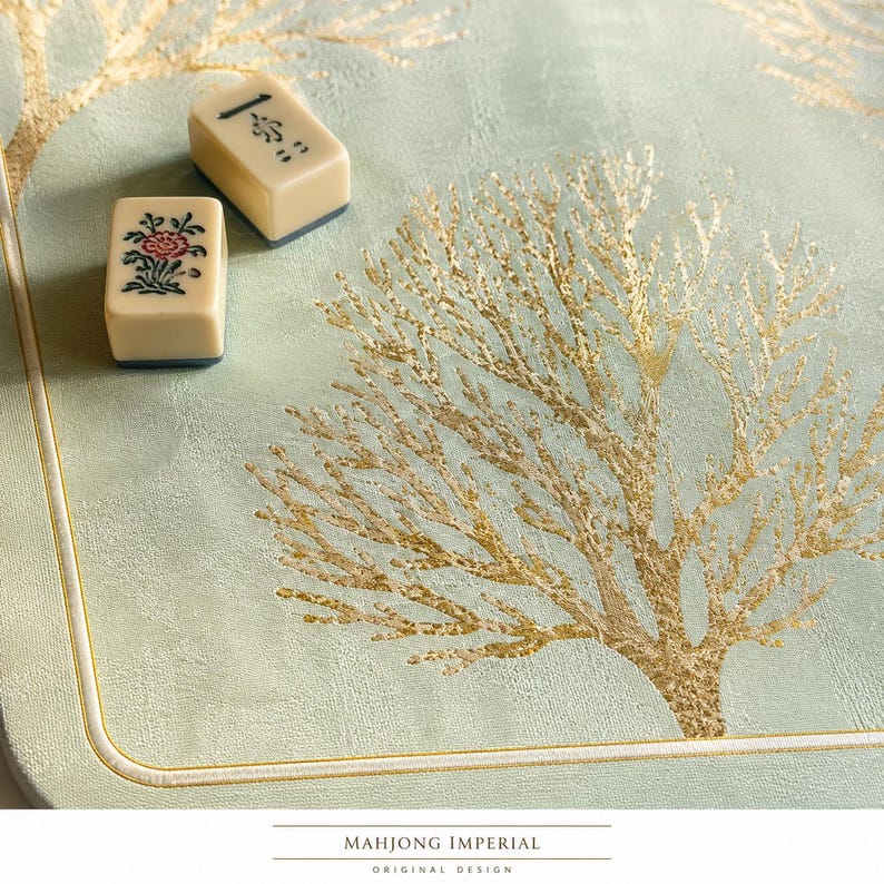 May include: A light green mat featuring a gold foil tree pattern and a white border, accompanied by two off-white mahjong tiles. One tile displays a black character, while the other shows a red and green floral design. Text reads MAHJONG IMPERIAL.