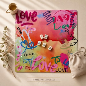 Könnte beinhalten: Eine quadratische Mahjong-Matte mit einem lebendigen, abstrakten Aquarellhintergrund in Rosa-, Orange- und Gelbtönen, überlagert mit dem Wort "love" in verschiedenen Schriftarten und Farben, dazu fünf Mahjong-Spielsteine und ein beiger Beutel.