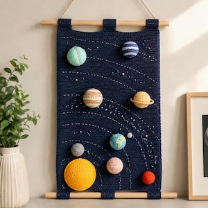 May include: A dark blue crocheted wall hanging featuring a solar system design with various colored, spherical, crocheted planets and a large yellow sun attached to the fabric, suspended from two horizontal wooden dowels with a string.