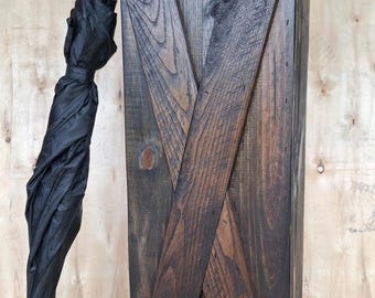 May include: A dark brown wooden umbrella stand featuring a prominent X-shaped cross brace design on the front panel. A closed black umbrella with a cylindrical handle is positioned vertically against the side of the rectangular wooden stand.