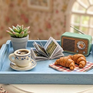 Könnte beinhalten: Ein hellblaues Holztablett enthält eine kleine Sukkulente im Topf, eine kleine Keramiktasse mit Kaffee auf einer Untertasse, ein offenes Miniaturbuch, ein mintgrünes Vintage-Radio und zwei Croissants auf einer karierten Serviette.