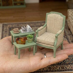 Puede incluir: Un conjunto de muebles de casa de muñecas en miniatura que incluye un sillón verde salvia con tapicería beige texturizada, dos mesas auxiliares a juego, una taza de café marrón, un libro abierto, una pequeña suculenta y una radio vintage.