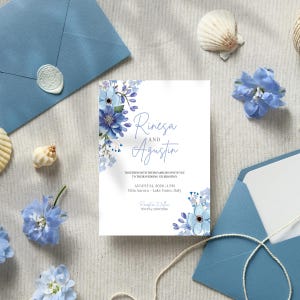 May include: A white wedding invitation card featuring blue floral illustrations, with the text Rinesa AND Agustin, August 24, 2026, 4 PM, Villa Aurora, Lake Como, Italy, and RSVP by 20.07.2026, accompanied by blue envelopes and blue flowers.