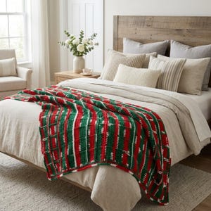 May include: A rectangular crochet throw blanket featuring a repeating vertical striped pattern in alternating shades of forest green, bright red, and cream white. The textured knit fabric is draped across a bed with neutral-toned bedding.
