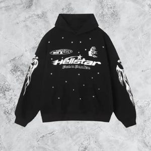 Puede incluir: Una sudadera con capucha negra con estampados gráficos en blanco. El pecho muestra el texto Hellstar con un icono de estrella, un logotipo ovalado más pequeño y una forma de llama. Ambas mangas tienen llamas blancas y el frente tiene estrellas.