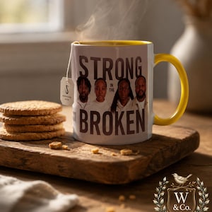 May include: A white ceramic mug with a yellow interior and handle, featuring a graphic of four people and the text "STRONG IN THE BROKEN". A tea bag with a tag labelled "S SCRIPTURE SAYS" hangs from the rim, and steam rises from the mug.