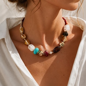 Peut inclure: Un collier de perles multicolores présentant une variété de formes et de textures, incluant des perles métalliques dorées polies, des pierres lisses blanches et translucides, une perle turquoise vive, une perle rouge foncé et une perle blanche.