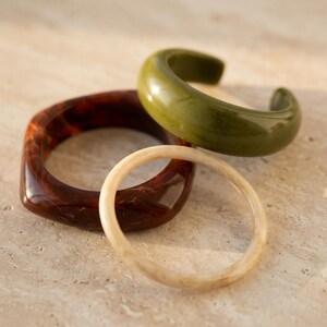 Può includere: Un set di tre bracciali composto da un polsino spesso verde oliva, un bracciale rigido largo con motivo tartarugato marrone scuro e un bracciale circolare sottile color crema. I bracciali presentano superfici lisce e lucide con bordi arrotondati.