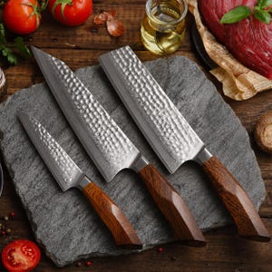 May include: A set of three kitchen knives with hammered metal blades and brown wooden handles. The set includes a small paring knife, a medium-sized chef knife, and a large rectangular cleaver, all featuring a distinct textured finish.