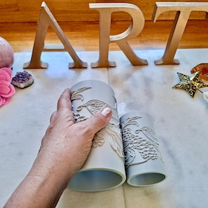 May include: Two cylindrical light grey silicone moulds featuring intricate embossed designs of birds and floral patterns. The larger mould is held by a hand, while the smaller mould sits beside it, both displaying detailed textures for crafting purposes.