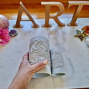 May include: Two cylindrical light grey candle sleeves featuring an embossed dragon design with intricate scale and wing details. The sleeves are positioned side-by-side, with one larger cylinder and one smaller cylinder, both displaying a textured pattern.