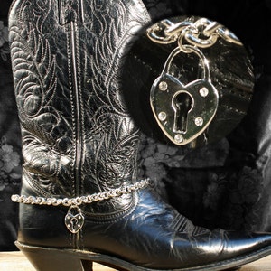 May include: A black leather cowboy boot with a silver chain ankle bracelet featuring a heart-shaped padlock with a keyhole and rhinestones. The text "Pamela Boot Anklet Jewelry" is written above the boot.