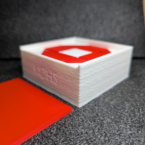 以下が含まれることがあります： 3Dプリントされた質感のある白い正方形の箱で、側面には「SHON」の文字が浮き彫りになっています。箱の中には、中央に白い正方形の切り抜きがある赤い正方形の物体が入っています。箱の横には、平らな赤い正方形の蓋が置かれています。