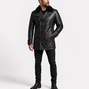 Op de afbeelding: Een zwarte, dubbelrijige leren jas met een donkere kraag van imitatiebont, lange mouwen met gespmanchetten en een knoopsluiting. De jas wordt gecombineerd met een zwarte broek en zwarte leren laarzen, wat zorgt voor een monochroom geheel.