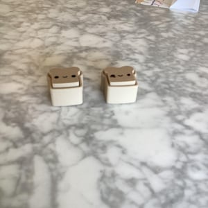 May include: Two small, rectangular, beige-colored plastic objects shaped like slices of toast. Each piece features a light brown top section with a printed face consisting of two black eyes and a small, curved mouth, resting on a solid beige base.