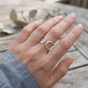 May include: A polished silver open-style ring featuring a smooth, rounded band that curves into two distinct, parallel U-shaped ends. The ring is worn on the middle finger, showcasing a minimalist, continuous metal design with a reflective finish.