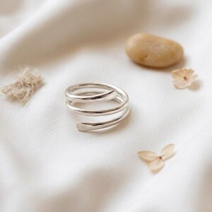 May include: A silver-colored ring featuring a triple-coil spiral design with a smooth, polished finish. The band wraps around itself three times, creating an open, layered appearance. The metal surface reflects light, highlighting the rounded edges.