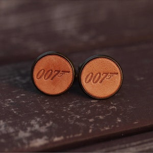 Op de afbeelding: Een paar manchetknopen van bruin leer met een gouden metalen frame. De manchetknopen hebben het nummer "007" gegraveerd op elk.