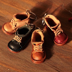 May include: Four miniature leather boot keychains in various colors: tan, black, brown, and burgundy. Each boot has laces and a metal keyring. The boots are arranged on a wooden surface.